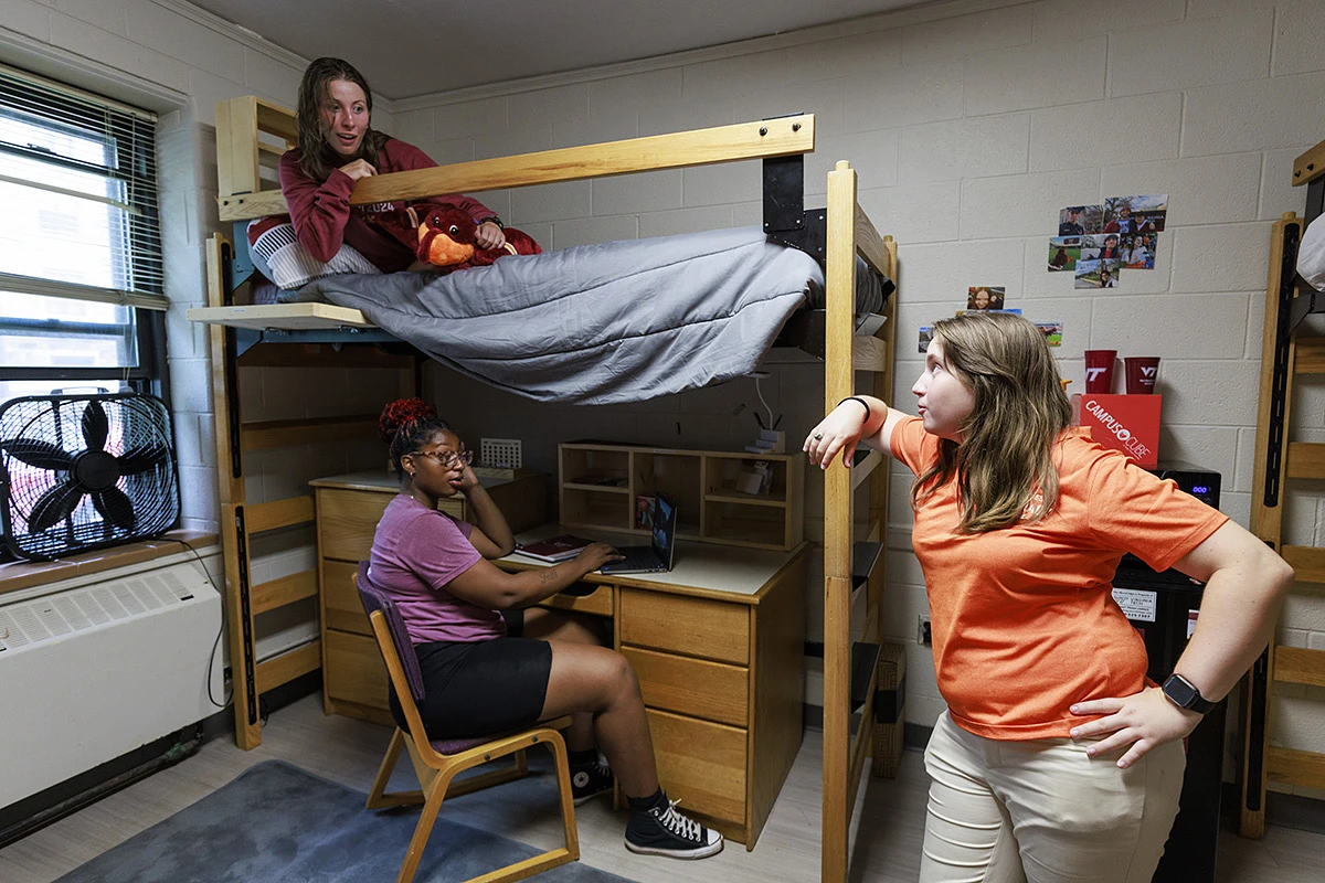 Three people are gathered in a Virginia Tech residence hall room, with one sitting at a desk, another on a lofted bed, and the third standing nearby.