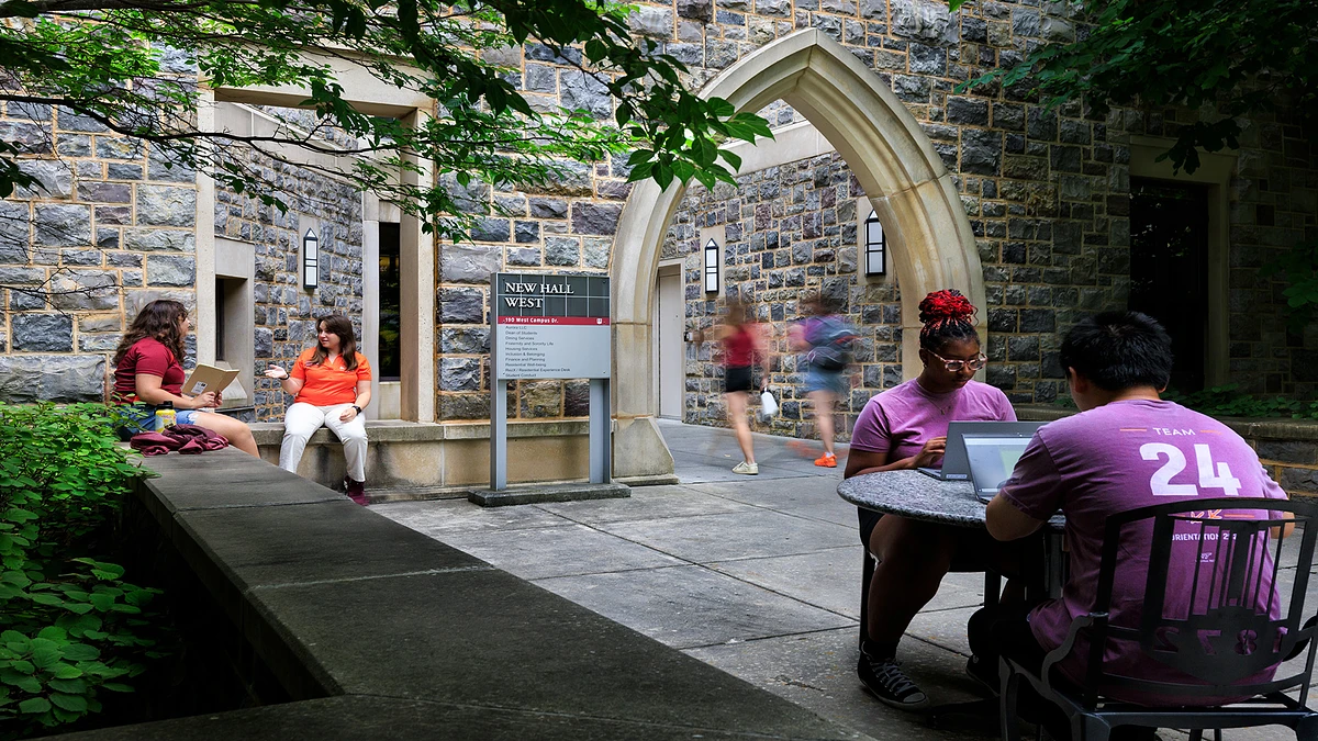 Students are gathered outside a hokie stone building, working on laptops and conversing near an arched entrance labeled "New Hall West."