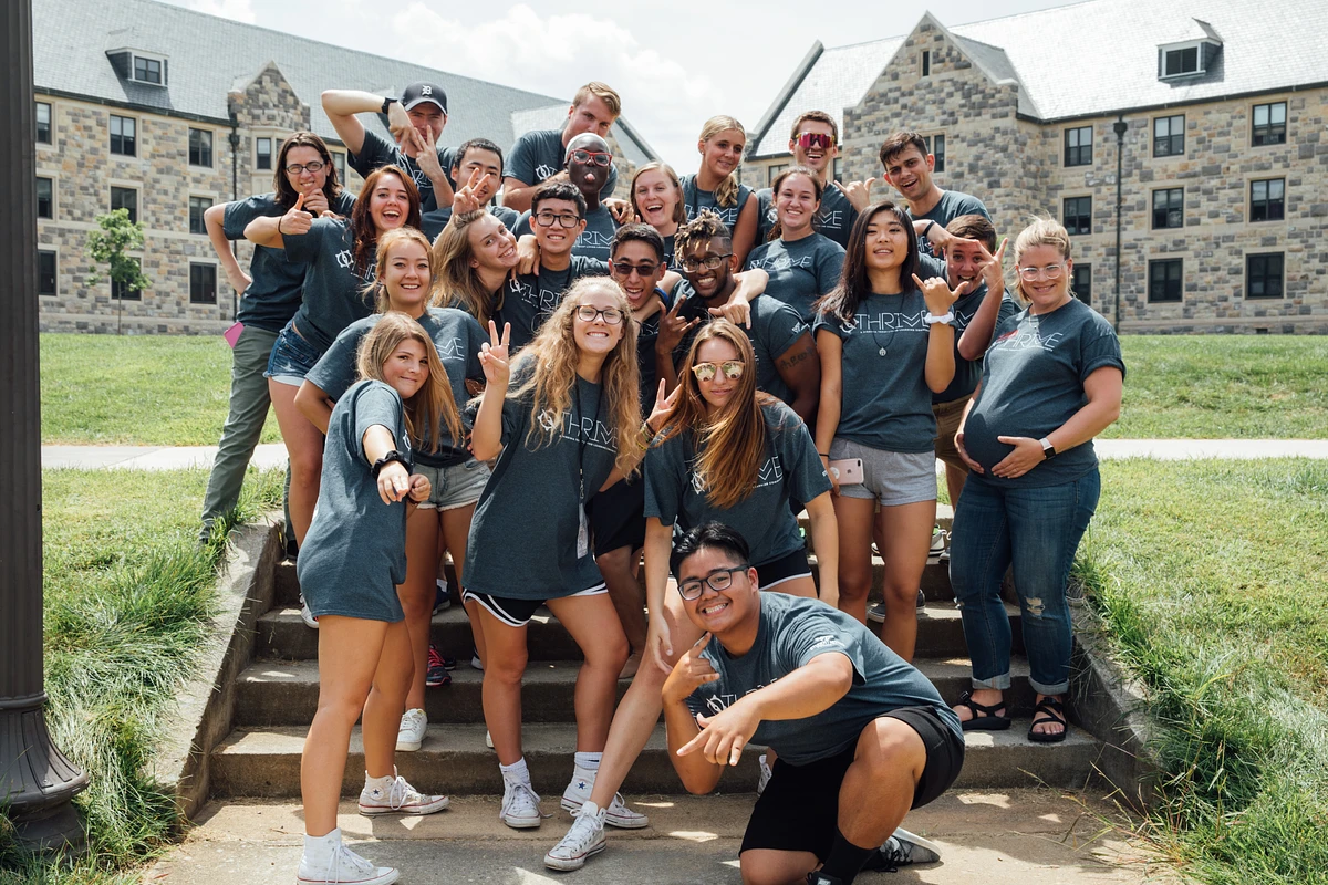 A group of people wearing matching "THRIVE" shirts pose energetically outdoors in front of a hokie stone residence hall.