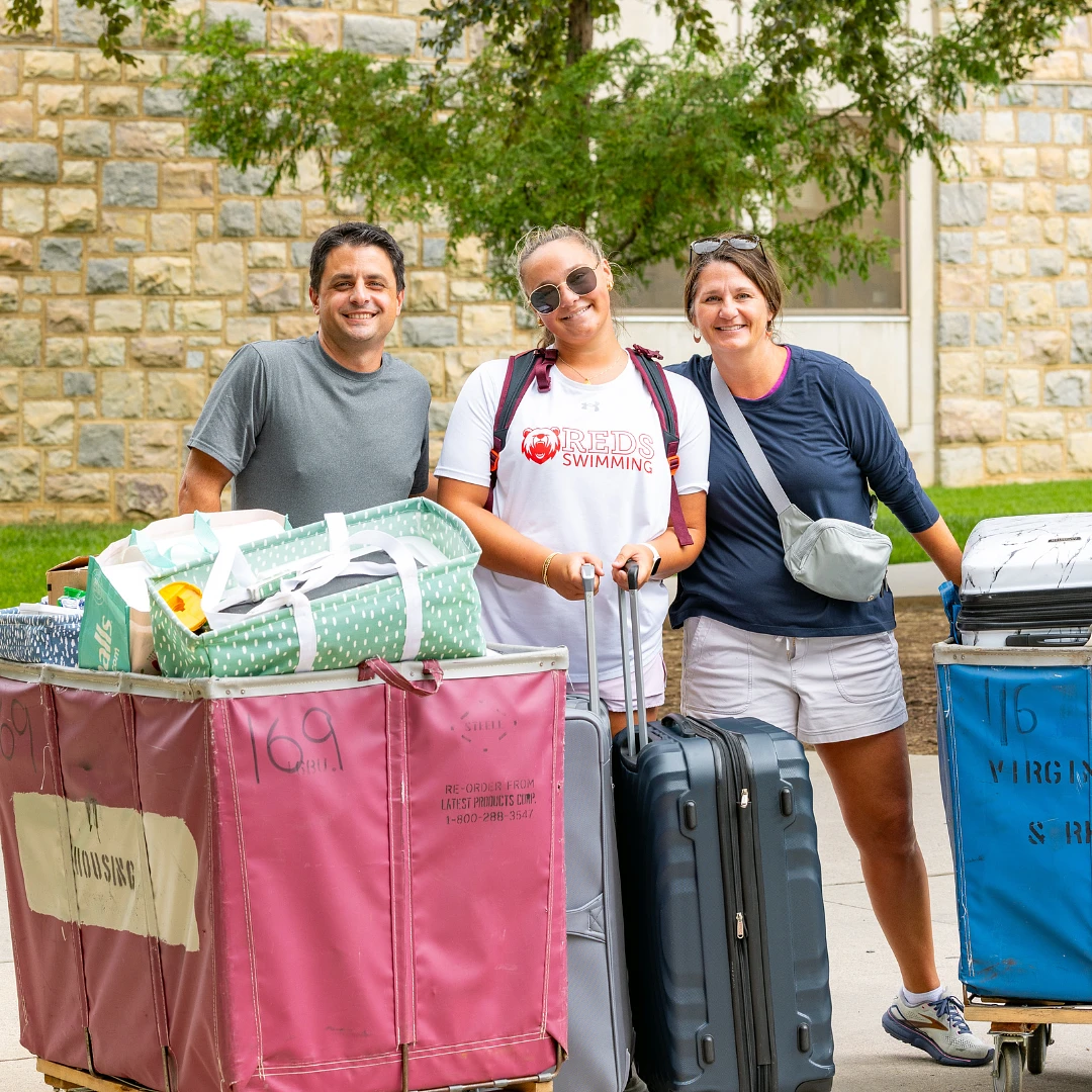 Three people pose cheerfully with luggage by large carts labeled for housing, possibly during a college move-in day.