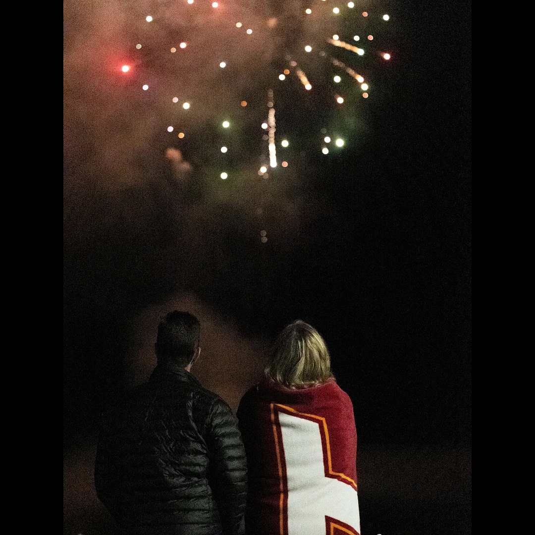 Two people stand closely together, watching fireworks light up the night sky. One is wrapped in a maroon VT blanket.