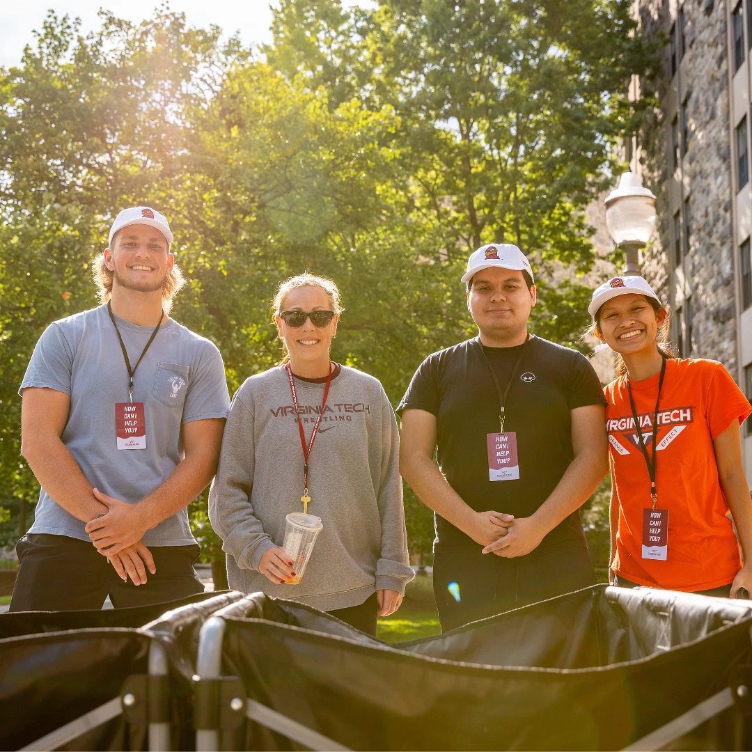 Four people wearing Virginia Tech apparel and "How Can I Help You?" tags pose together outdoors on a sunny day.