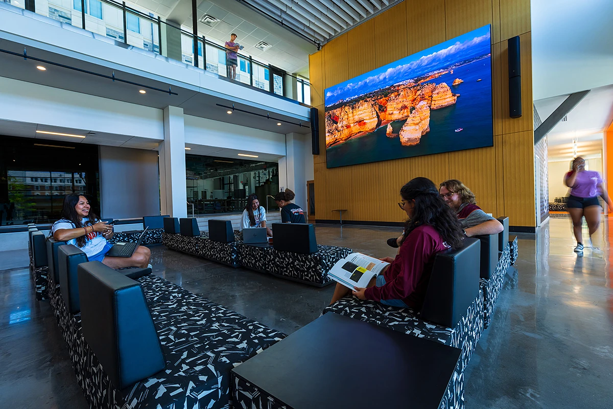 People are socializing and studying on assorted couches in a modern, open space with a large digital display on the wall.
