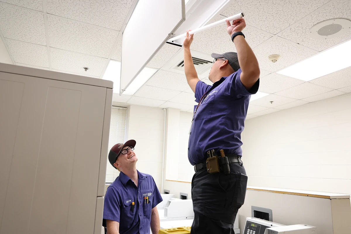 Two workers in uniform are changing a fluorescent light tube in a laundry room ceiling.