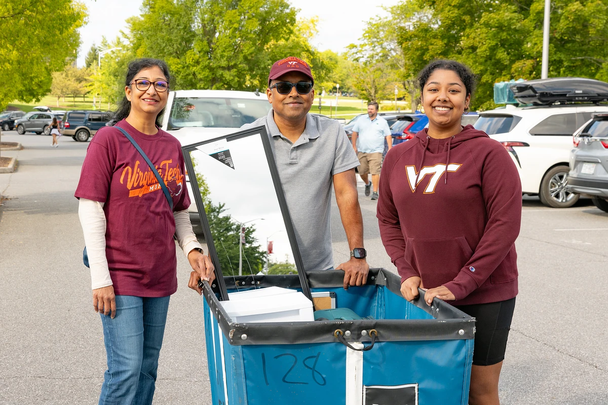 Three people are smiling and standing outdoors next to a cart filled with items, with a parking lot and trees in the background.