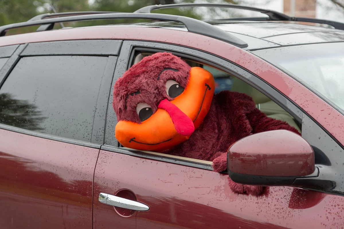 A person in a large maroon mascot costume leans out of the window of a similarly colored car.